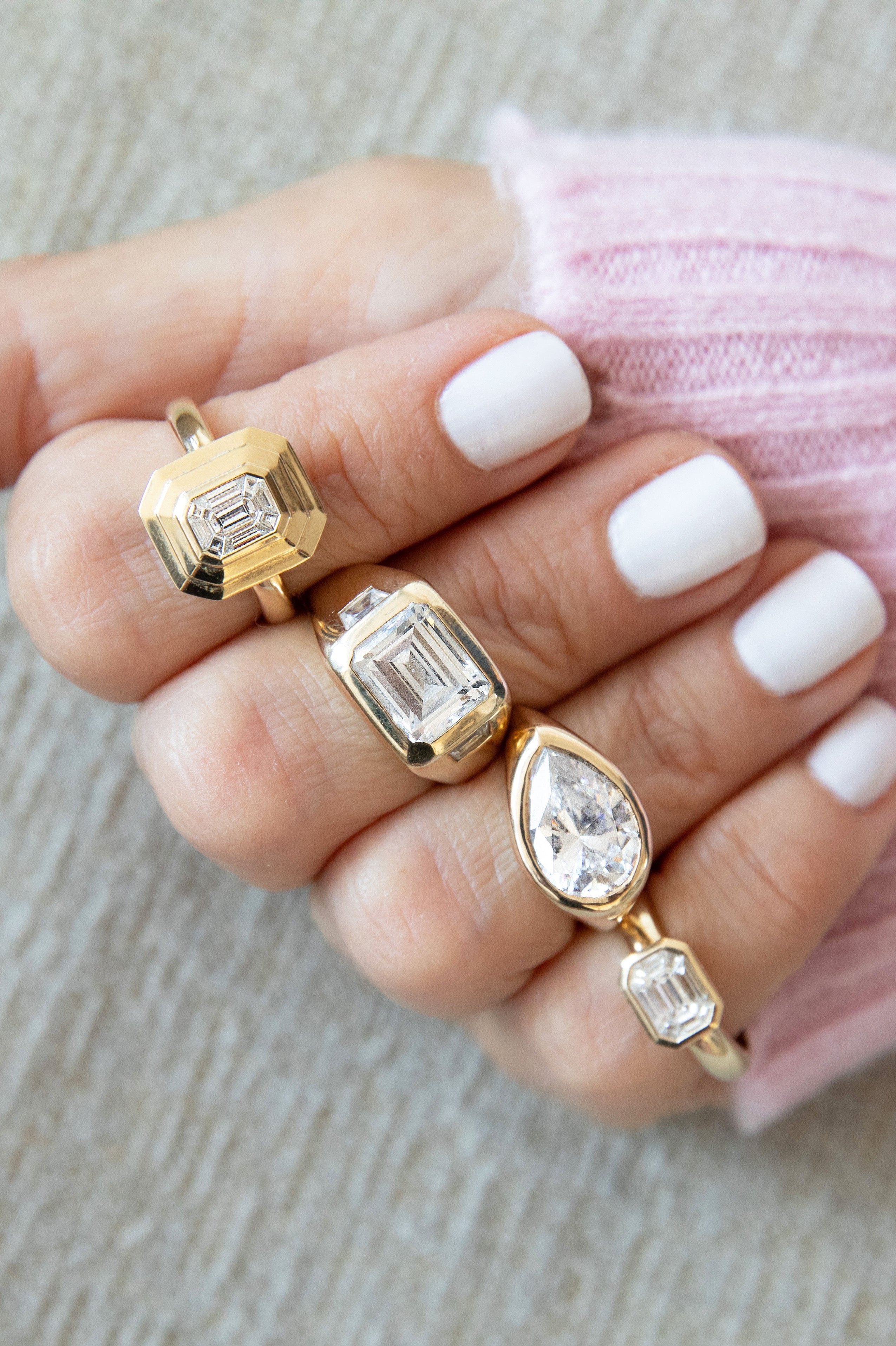Hand with four gold rings, each featuring a large diamond in different cuts: an emerald, a cushion, and a pear-shaped.