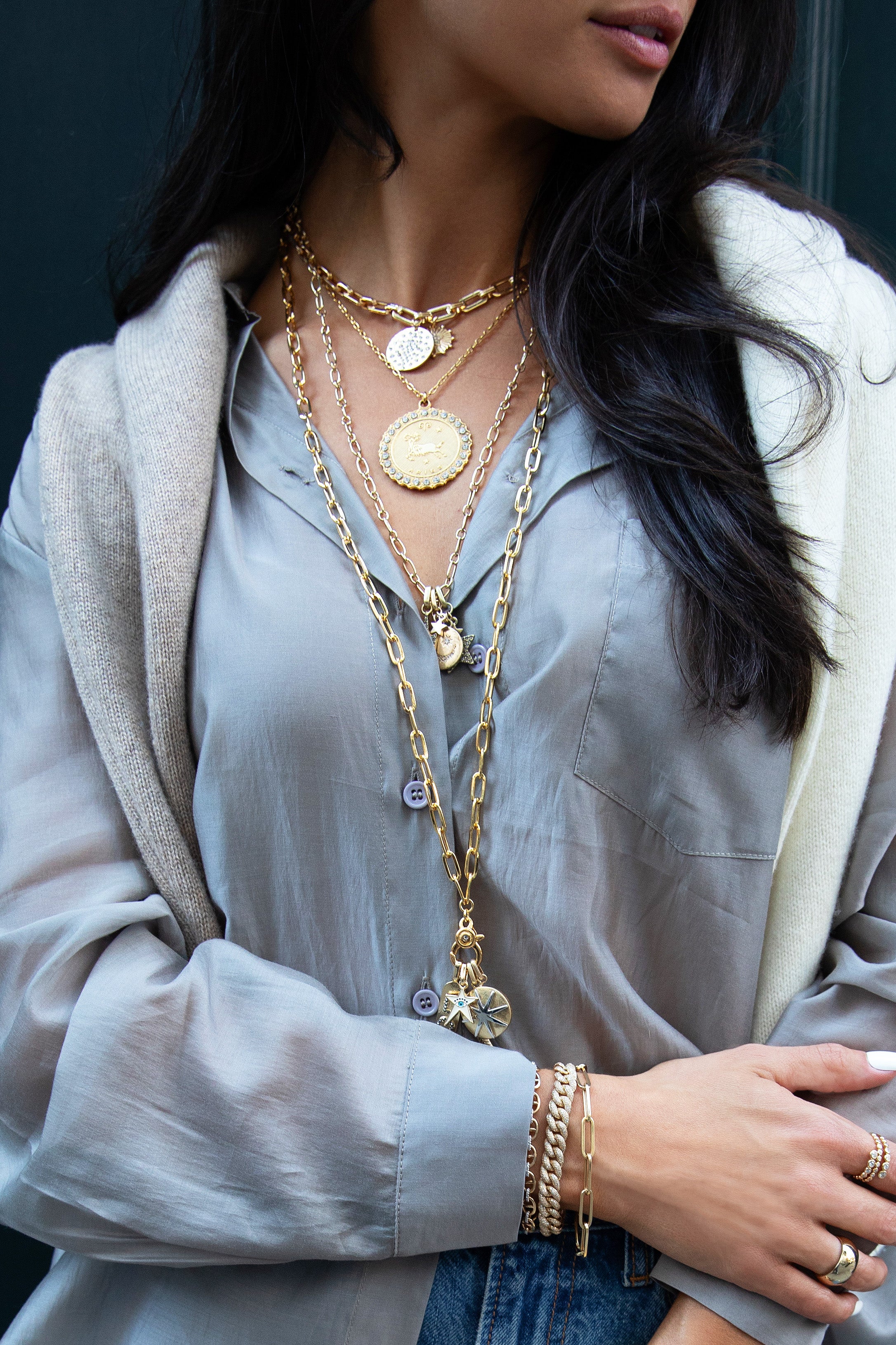 Close-up of a person's torso and arm, showing a grey shirt, a beige sweater draped over the shoulders, and multiple layered gold necklaces with various pendants. The visible hand is adorned with several bracelets and rings.