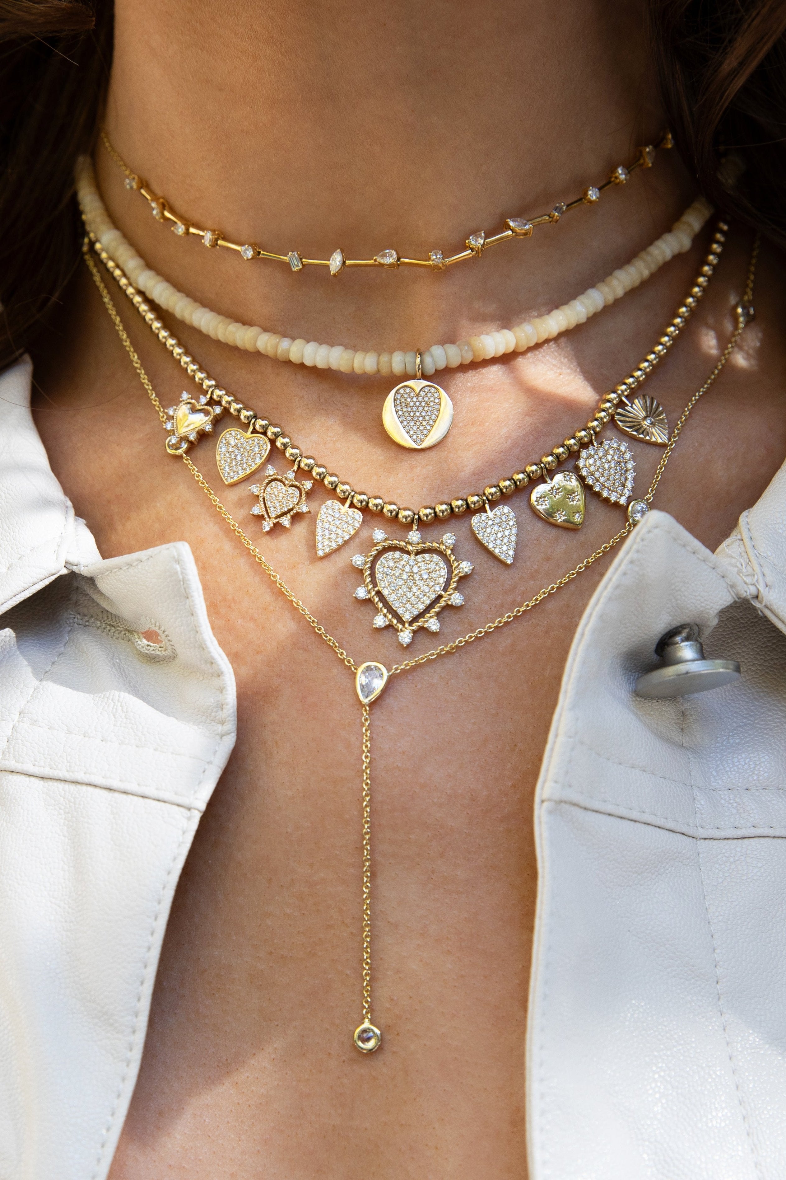 A neck party featuring delicate gold chain with small diamonds, a choker made of light-colored beads with a heart pendant, and several gold chains adorned with various heart-shaped and other diamond-encrusted charms.