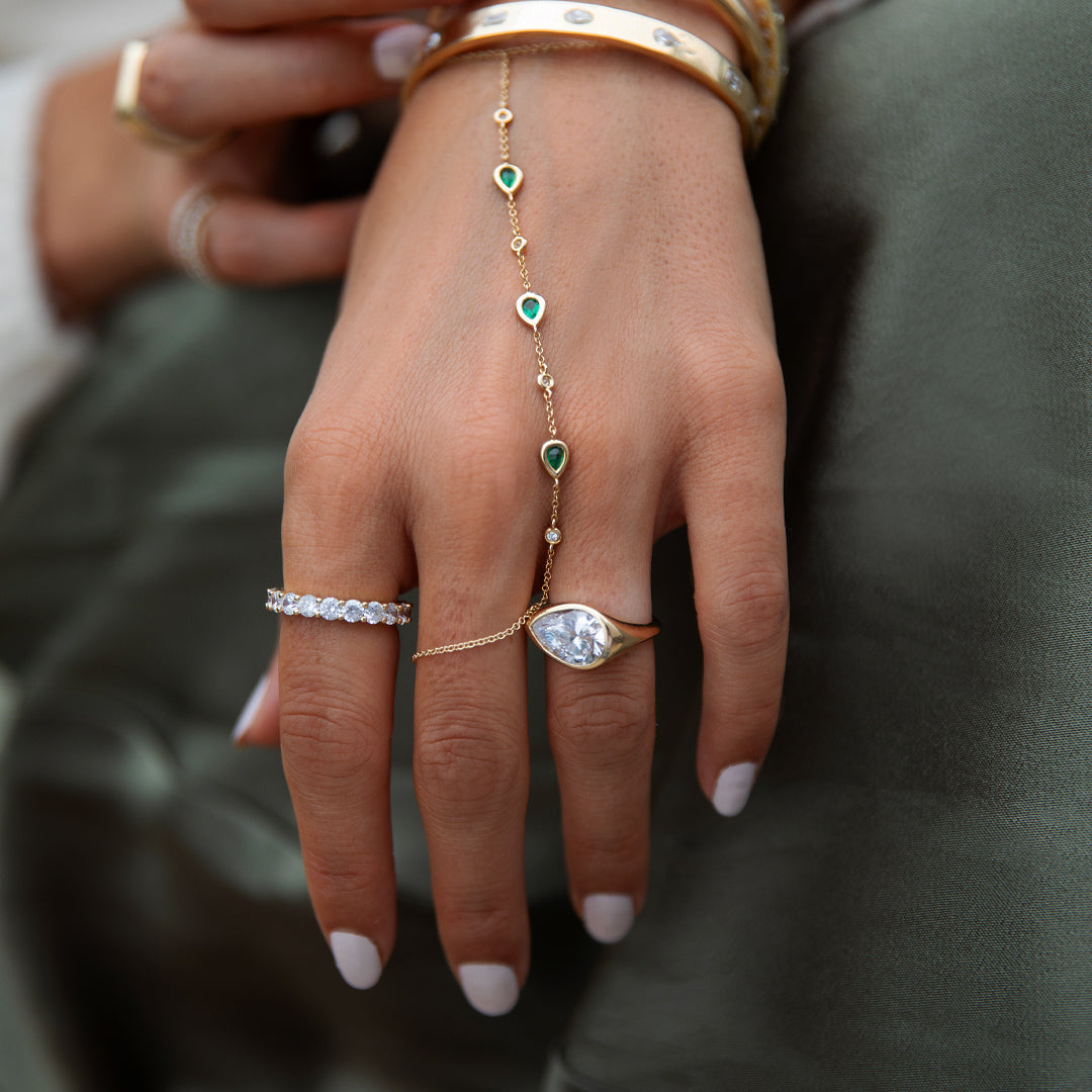 Hand wearing multiple gold and silver rings with emerald and diamond stones on a dark background