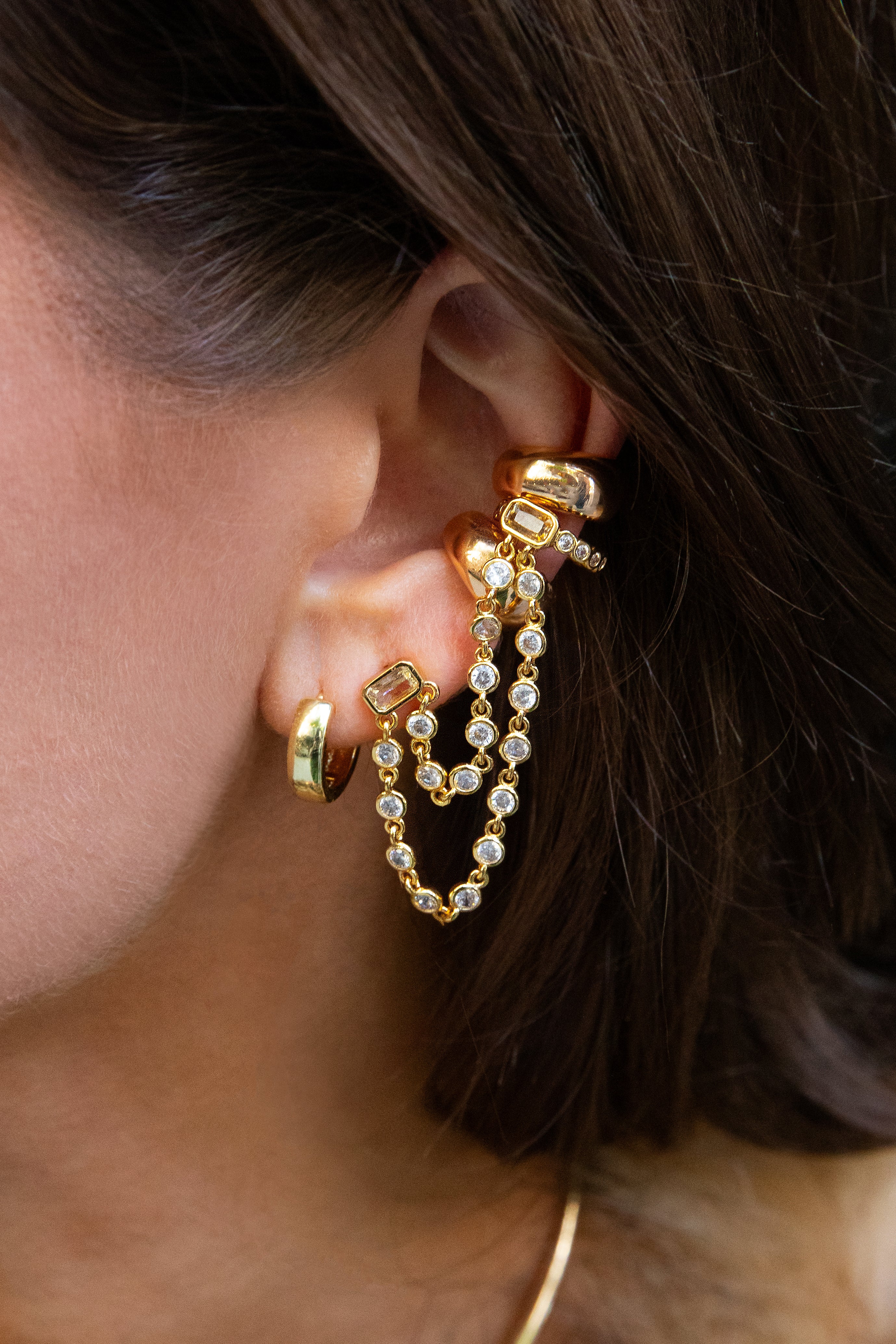 Close-up of a woman's ear wearing layered gold earrings, including a statement chain with sparkling stones connecting the lobe to the upper ear.