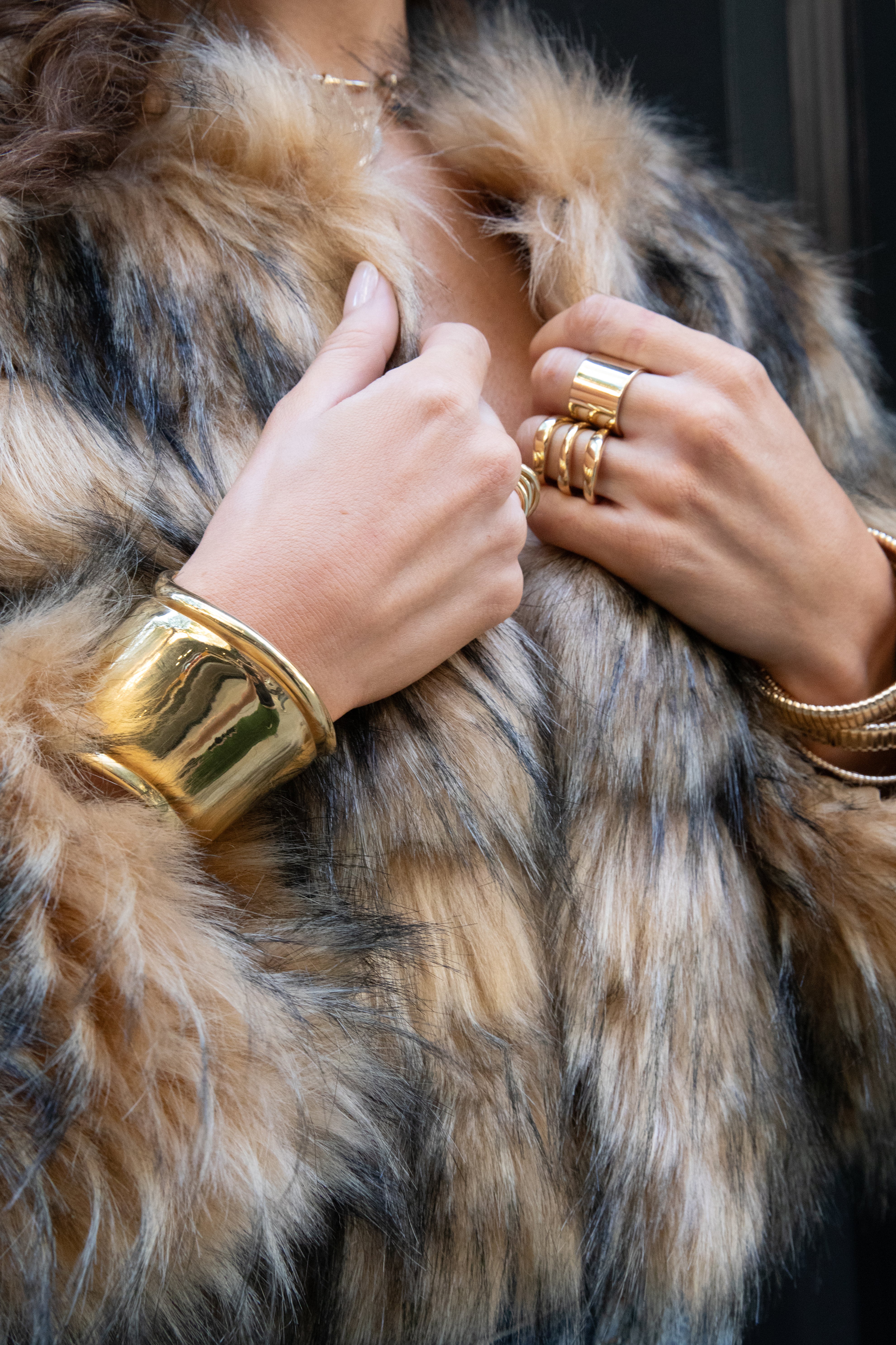 A person wearing a textured brown and black faux fur coat, gold rings and a large gold cuff bracelet.