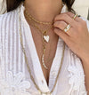 White enamel heart charm displayed on yellow gold chain link lariat, worn layered with gold chains and with chunky gold rings