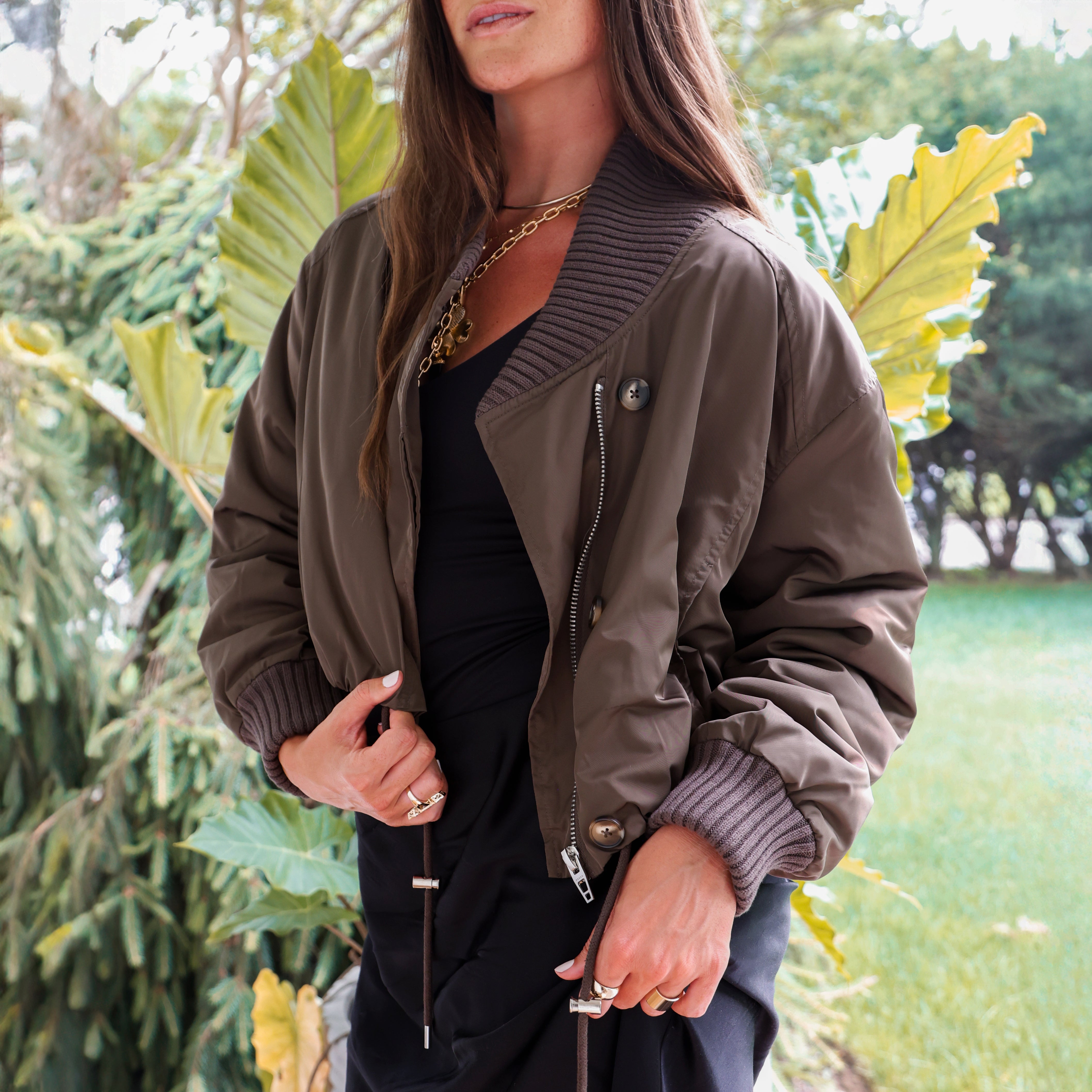A woman wearing an olive brown bomber jacket with ribbed knit collar, cuffs, and hem, styled over a black top in an outdoor garden setting.