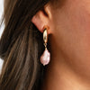 These exquisite hoops feature a stunning light pink pearl elegantly set in your choice of yellow or white gold plating, embodying the essence of elegance and hope. These gorgeous earrings perfectly complement any ensemble, making them a versatile addition to your jewelry collection. Pair them effortlessly with other jewels and stay in vogue while making a meaningful impact.   Pink Pearl Drop on Small Hoop Pieced Earrings  Yellow or White Gold Plating