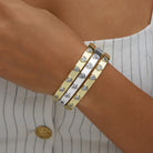 A close-up of a person's wrist wearing three slim metallic bangles. The bangles are gold and silver with small clear heart-shaped stones set along the front.