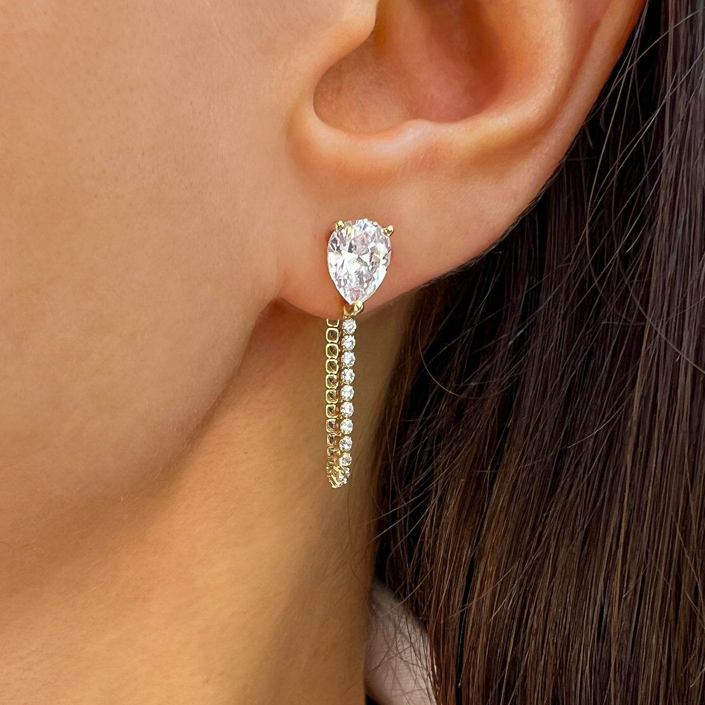 Close-up of a woman's ear wearing a gold teardrop earring with a central gemstone and dangling chain.