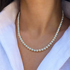 A close-up of a gold tennis necklace with clear round-cut stones worn by a model in a white collared shirt.